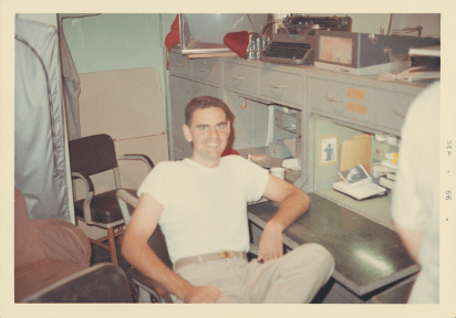 Colored image of a man seated in a chair smiling at the camera with his left arm resting on a desk, a typewriter is on the shelf above his head