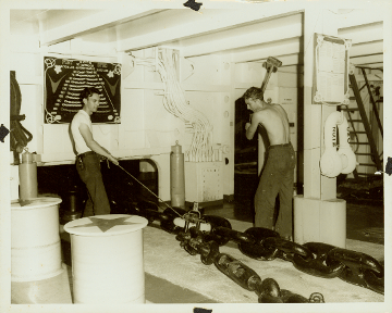 Black and white photo of two men on either side of anchor chain, one man holds rope attached to chain and other raises a large hammer 