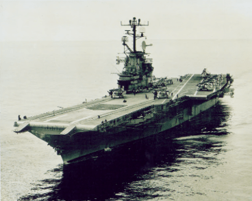 Black and white aerial photo of aircraft carrier USS Intrepid at sea, crew members and airplanes visible parked on aft flight deck 