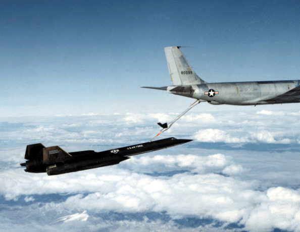  A black A-12 jet flies over clouds; a fuel line running from its top is connected to back of a larger grey jet plane that is partially visible above it.