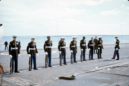 Color photo of eight Marines wearing dress uniforms standing in a line on the flight deck of USS Intrepid