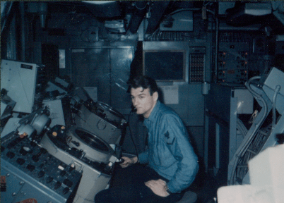 Color photo of a man wearing blue work uniform seated in front of radar equipment looking at the camera