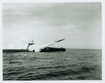 Black and white image of USS Growler above water launching Regulus I missile into the air, cloud trail extends behind missile partially obscuring submarine