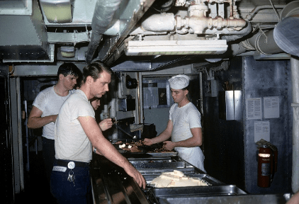 Color photo of three men serving themselves food from metal chafing dishes, another man stands behind the cafeteria line wearing an apron