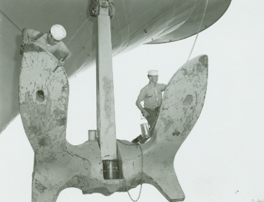 Black and white photo of large hanging anchor with two sailors on each side of the anchor, scraping paint