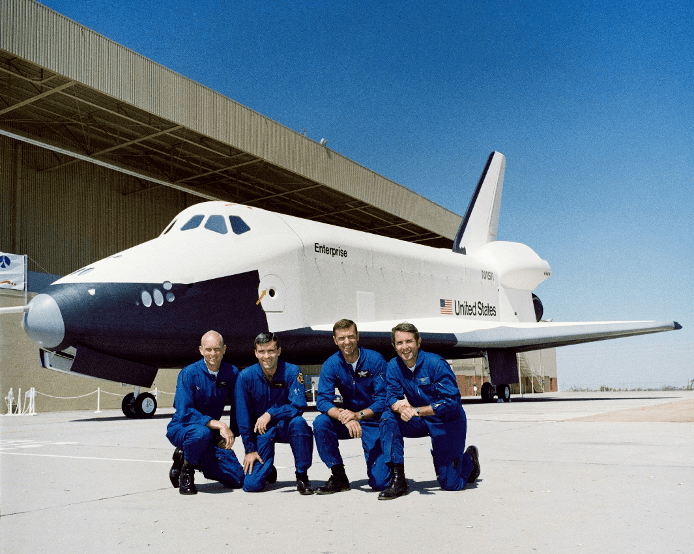 The four test pilots in their blue flight suits are kneeling in front of the space shuttle orbiter Enterprise on an airport tarmac