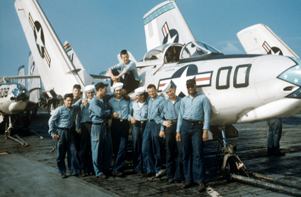 Color image of ten men posed in front of a Grumman F9F-6 Cougar airplane on the flight deck with wings folded up, other airplanes visible in background