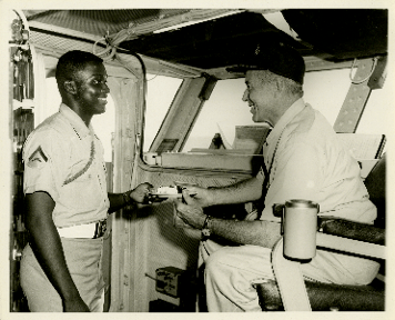 Black and white image of dark-skinned man handing a plate with a piece of cake to a light-skinned man seated in a chair