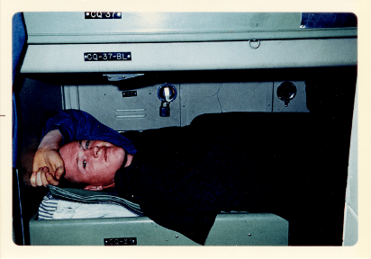 Color photo of man lying in berthing bunk with left forearm resting on his forehead, locker visible behind him