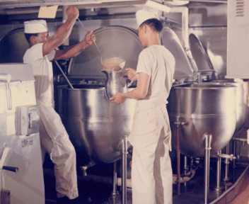 Color photo of two men, wearing all white uniforms, standing in front of a large silver kettle, using ladle to transfer gravy to a metal pitcher