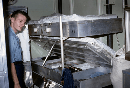 Color image of sailor standing next to bunk, the middle bunk is open to reveal a locker and storage compartments below