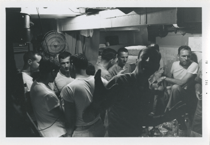 Black and white image of nine men inside a stateroom, they are standing or seated in circles talking, one man poses and smiles for the camera