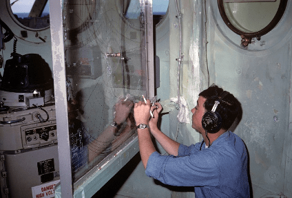 Colored image of a man wearing headphones kneeling down to write on a clear glass board