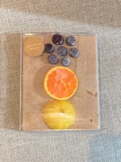  The following artifact is a touch object consisting of three types of fruits, each manipulated to form the the shape of a circle: seven whole blueberries, one cut cara cara novel orange, and one cut lemon. 