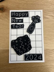 A white paper grid with embroidered objects placed on top; one reads "Happy New Year", another "2024", one is a champagne bottle being popped, and the last is confetti. 