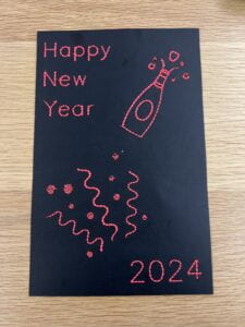 Black card with embroidered objects, one reads "Happy New Year", another "2024", one is a champagne bottle being popped, and the last is confetti in pink thread.