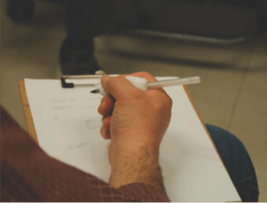 Man's hand holding a clipboard and a pen with a 3D printed customized grip. 