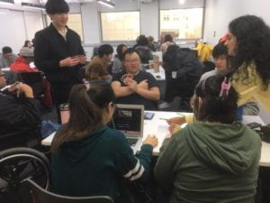 Students and Community members sitting at a table in a crowded classroom playing cards and working on a laptop.