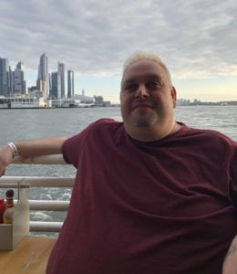 Gus Chalkias wearing a maroon shirt with the East River and Brooklyn skyline in the background. 