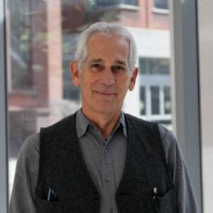 Headshot of Allan Goldstein in a gray shirt with black vest in front of a window. 