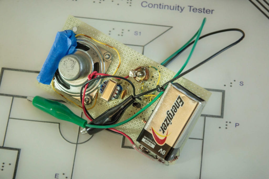 Close up of the continuity tester built in the workshop laying on top of a tactile schematic, which shows all connected components on a circuit board including a 9V battery, alligator clips, an 8 ohm speaker, and transistor.