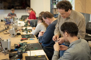 Six attendees sit at a long conference table at a blind soldering workshop, set up with individual soldering stations that include soldering mats, fans, helping hands, tools, a tactile schematic, and soldering irons. In the foreground, Josh Miele hands a circuit board to an attendee.