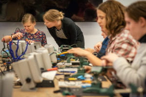 At the end of a conference table, an attendee focuses and holds their soldering iron, while a volunteer observes.