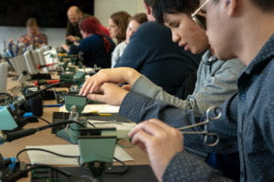 A long conference table, full of attendees, is set up with individual soldering stations with soldering irons, fans, vices, and soldering mats. In the foreground one attendee guides another's hand across their circuit board held by a vice at their soldering station.