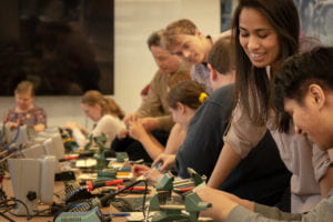 Two volunteers and Josh Miele lean over the shoulders of attendees—seated at their soldering stations at a long conference table—smiling and assisting.