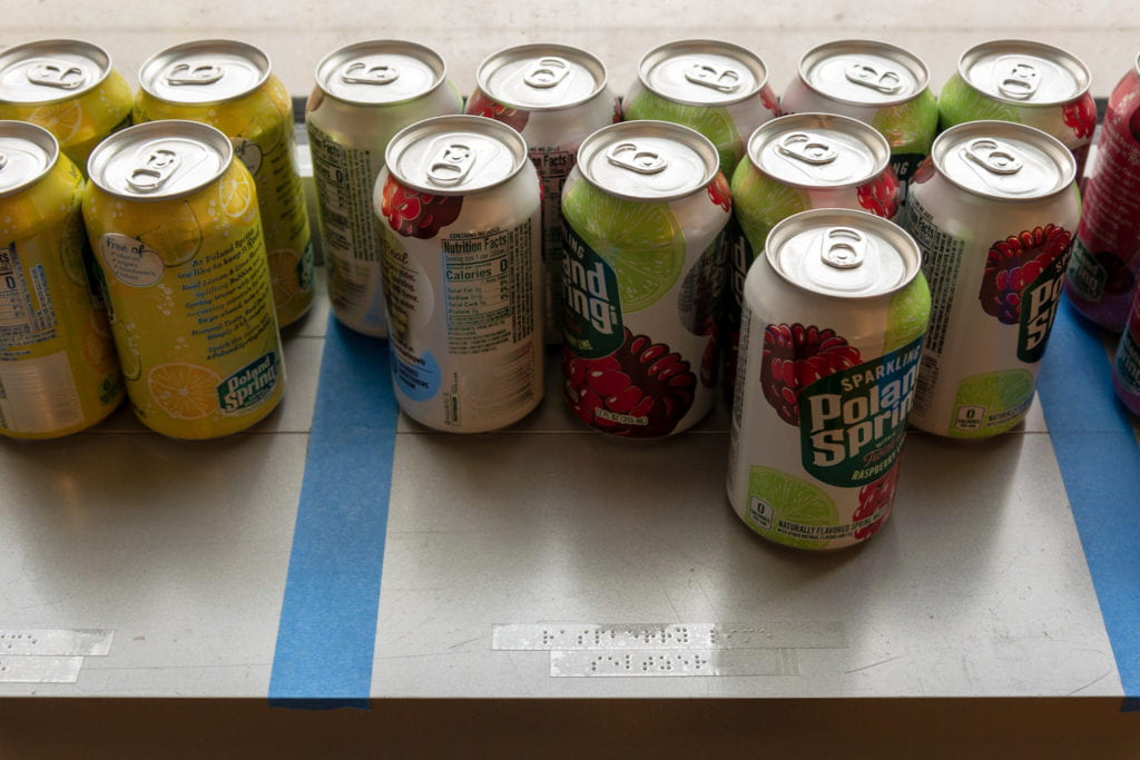 Cans of seltzer water on a window ledge, their flavors separated by parallel strips of gaffer tape. Towards the front of the ledge are adhesive Braille labels indicating the seltzer flavor.