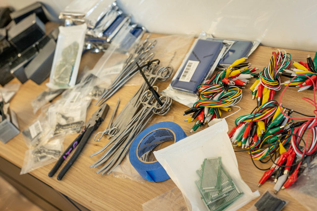 A cornucopia of tools and components used during the workshop lay across a desk, some steal sealed in their packaging, including, forceps, gaffer tape, enclosures, circuit boards, and alligator clips.