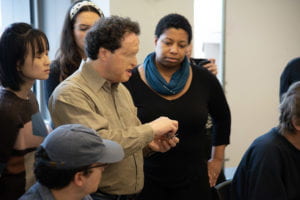 Volunteers gather around Josh Miele and listen intently as he demonstrates a step in building a continuity tester.