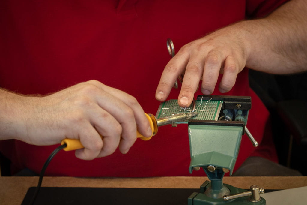 Close up of an attendee soldering their circuit board, secured in a vice. Their left middle finger touches the base of the forceps, which landmark where to line up the soldering iron tip.