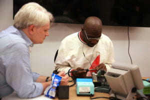 Tom Igoe watches an attendee who is concentrating intently on soldering at their soldering station. Delicate plumes of solder smoke can be seen wafting up from the tip of the iron.