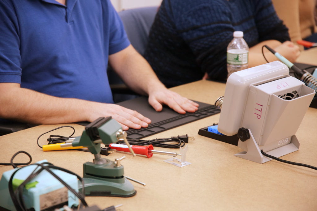 An attendee places their hands atop a bare soldering mat, with small trays spanning across the very top of the mat for small components. At their 12 o'clock is a fan and to their 2 o'clock is a soldering iron and vice.