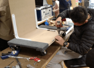 A person drilling pieces of plywood to a four-wheeled dolly. 