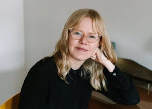 Lauren Race sitting at a desk wearing a black shirt and pink glasses, with her head propped up with her hand