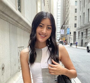 Viviana with black wavy long hair wearing white and grey patchwork sleeveless shirt is smiling while facing the camera