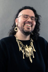 Head shot of Daniel Ryan Johnston from the waist up laughing with a black sweatshirt, gold chain necklace with plastic bones and pearls and matching earrings. 