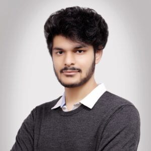 Brown man with dark hair and a light beard wearing a grey sweater over a white shirt.