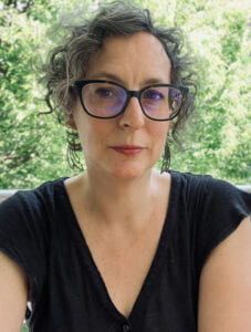 A white woman with grey-streaked curly short hair wearing glasses and a black shirt with bright green trees in the background. 