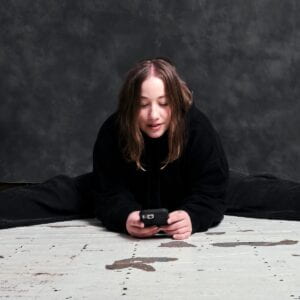 Blair sitting on the ground, holding a cellphone and laughing in front of a dappled grey photography background.