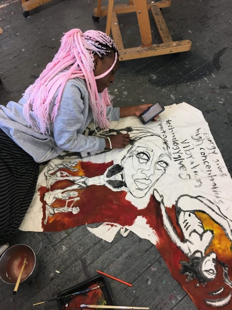 Student holding her phone in one hand and painting on canvas.