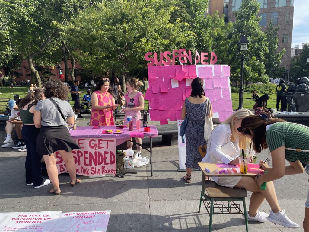 A public performance in a park. People dressed in pink with a large bulletin board saying 'Suspended'