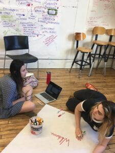 Two figures sit on the floor and work together on a large piece of paper with markers and a computer