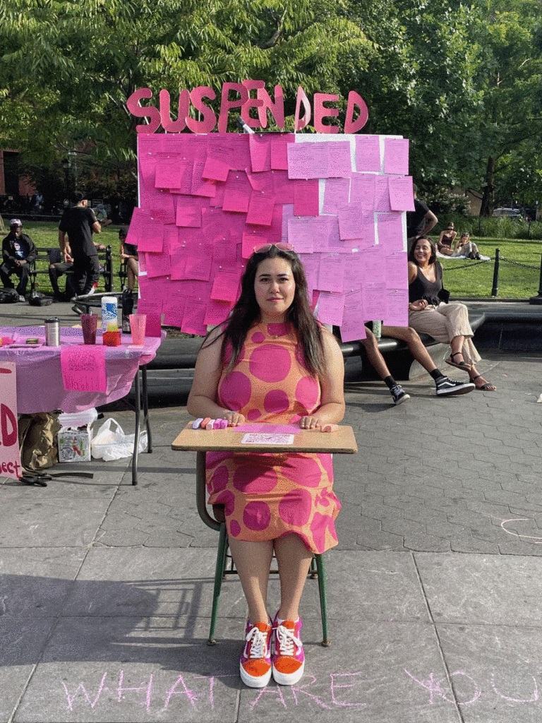 A Gif file includes 4 images of women dressed in pink sitting at a school desk in front of a bulletin board that says 'Suspended'