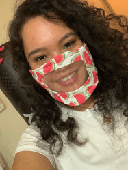 a close up picture of a girl with curly hair, smiling with teeth shown, and wearing a watermelon-printed face mask with a clear window in the middle of the mask 