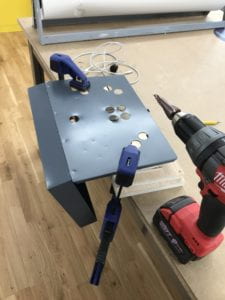 Clamped metal closure on two jigs with a screwdriver on a table next to a screwdriver