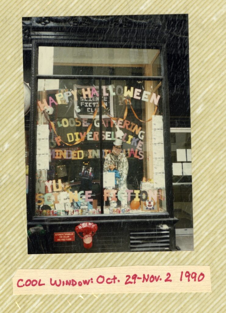 Image of a large, floor to ceiling window taken from the outside of a black building. The window has been decorated by the NYU Sci-Fi Club for Halloween. In colorful letters that have been cut out of paper are the words "Happy Halloween! A Loose Gather of Diversely Like-Minded Individiuals. NYU Science Fiction." The window is decorated with orange streamers, cans of soda, a cardboard cut out of a person, cans of soda, illegible flyers, and an image of a clown.