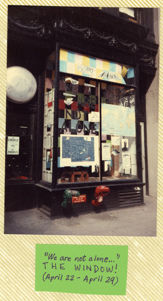 Image of the same window this time decorated with the words "The Science Fiction Club. We are Not Alone: A Festival of Foreign Science Fiction Films" cut out of black paper. Behind the letters are full sheets of colorful paper arranged in a checkerboard pattern, alternating between paper and blank spaces. There are 2 maps under the words. One map is a star chart with UFOs among the stars and the other is a map of the world. There are several unidentifiable black and white photographs and flyers hung around the maps.